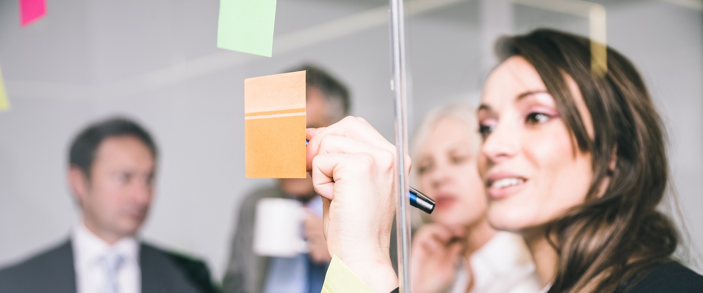 A business woman writing on a post-it while her colleagues talk behind her